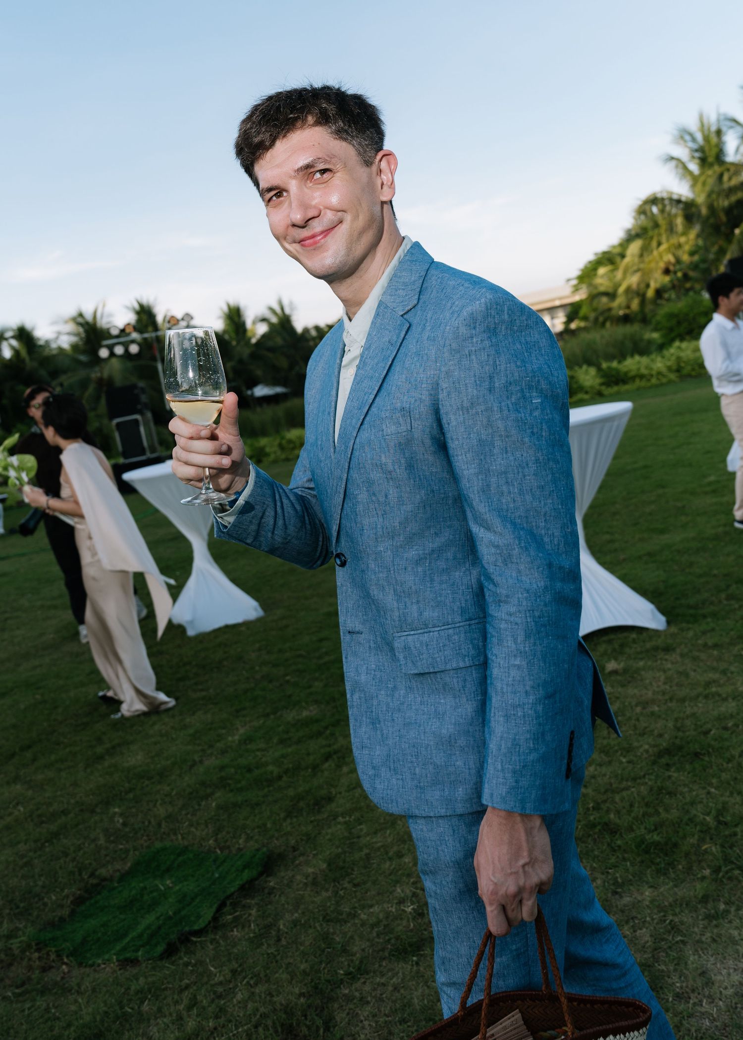 Smiling man in a light blue linen suit toasting with champagne at an outdoor garden wedding reception.