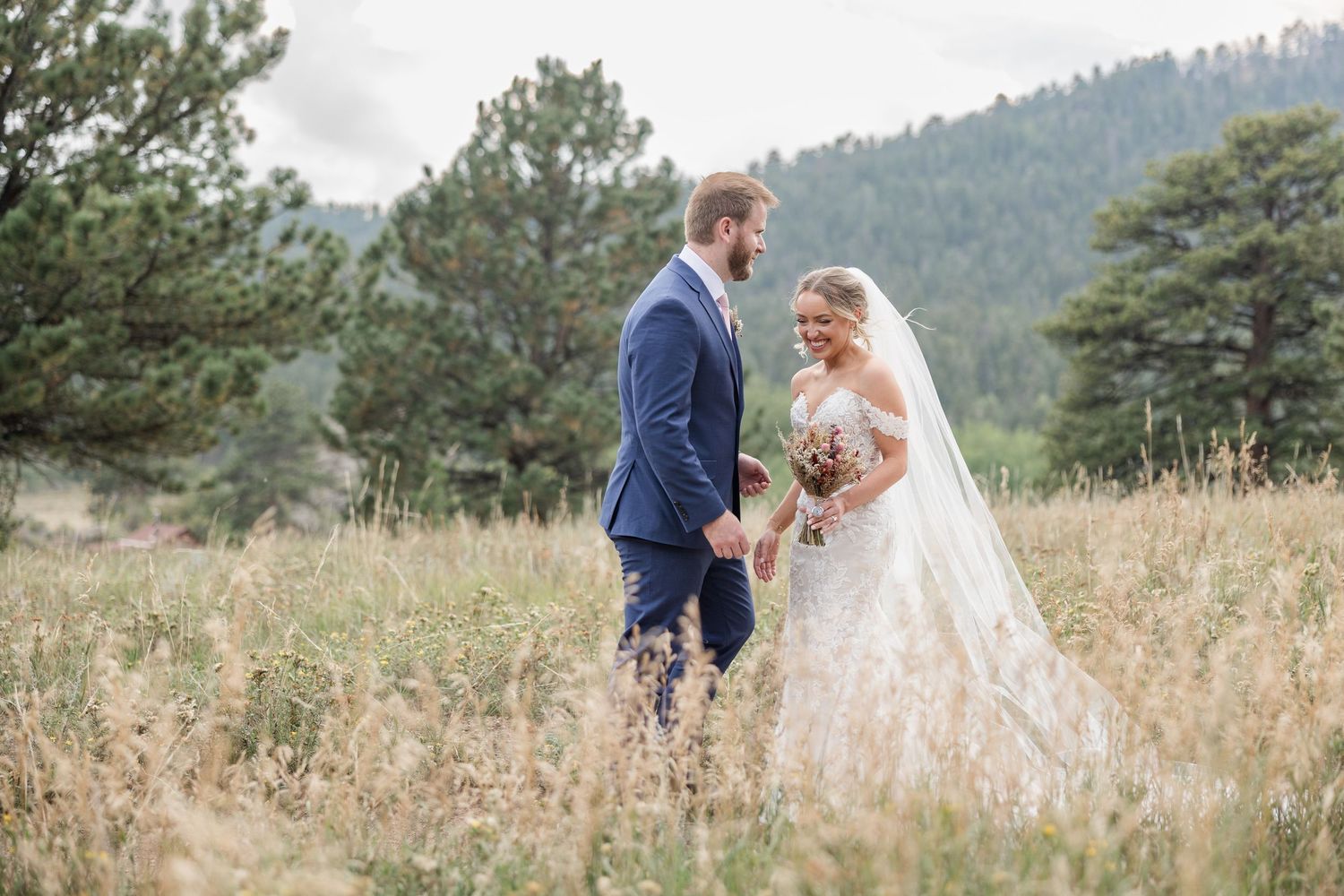 Newlywed couple shares romantic moments in a scenic mountain meadow with tall grass and evergreen trees.