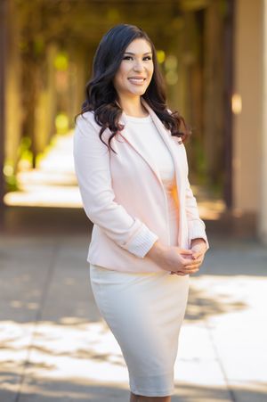 Professional portrait of a business woman in a light pink blazer and white dress smiling outdoors in natural lighting.
