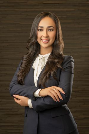 Professional headshot of a woman in a dark business suit and white tie-neck blouse against a textured background.