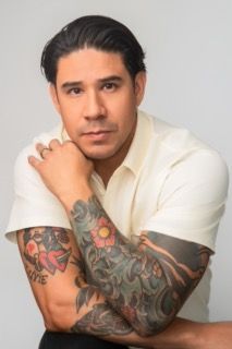 Portrait of someone wearing a white shirt showing colorful tattoo sleeve artwork while posing with hand near chin.
