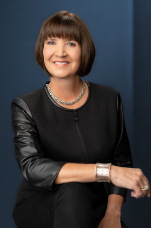 Professional business portrait of a person wearing a black jacket with leather sleeves and silver jewelry against a blue background.