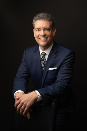 Professional corporate headshot of person in navy suit with striped tie against dark background.