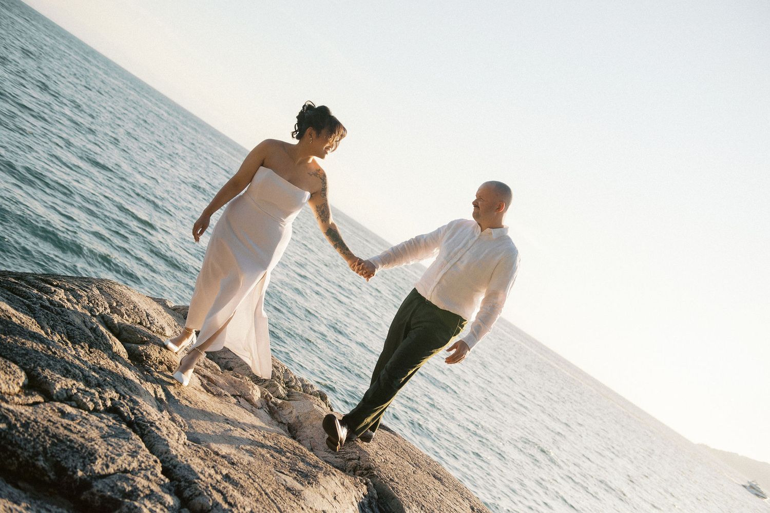 Couple walking hand in hand on rocky coastal outcrop with ocean view.