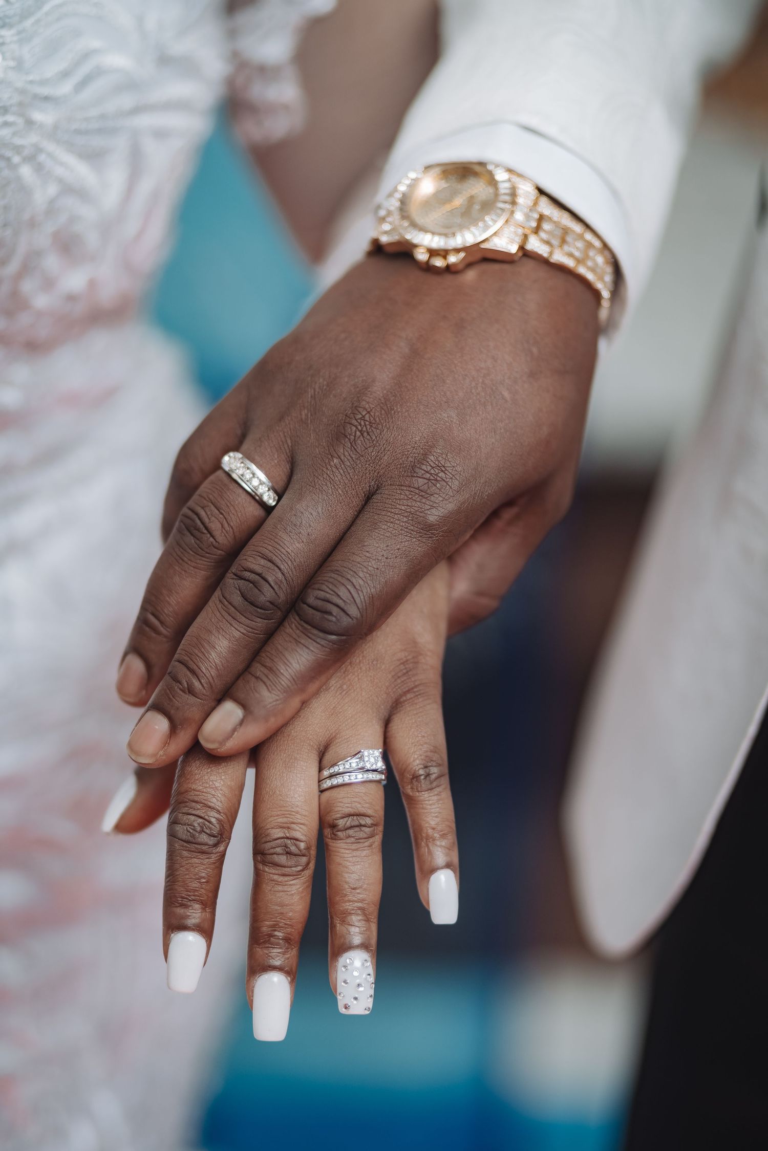 Close-up of hands wearing wedding rings and a gold watch, with white manicured nails against a blue background.