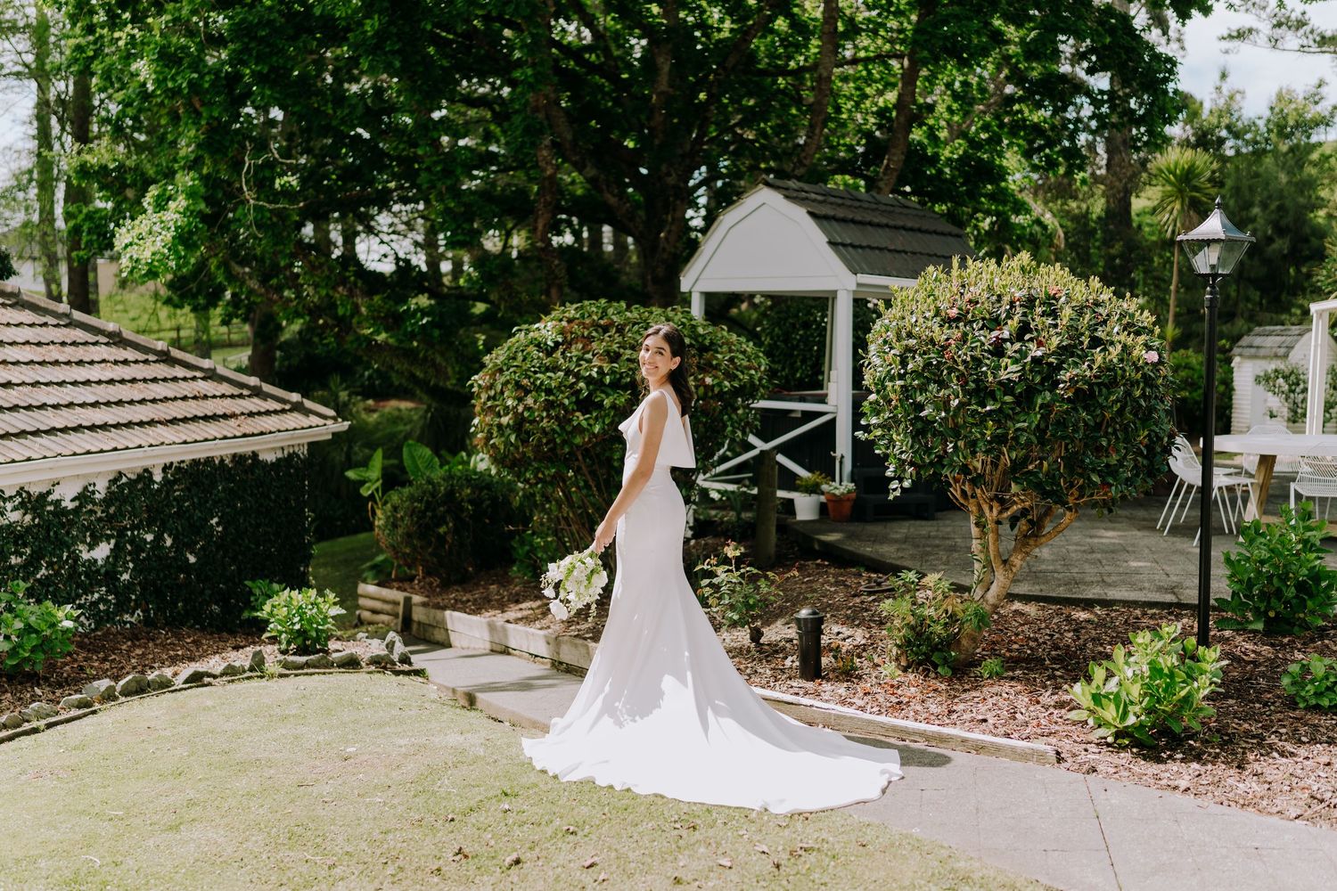 A long white wedding dress with a flowing train on display in a scenic garden setting with a white gazebo.