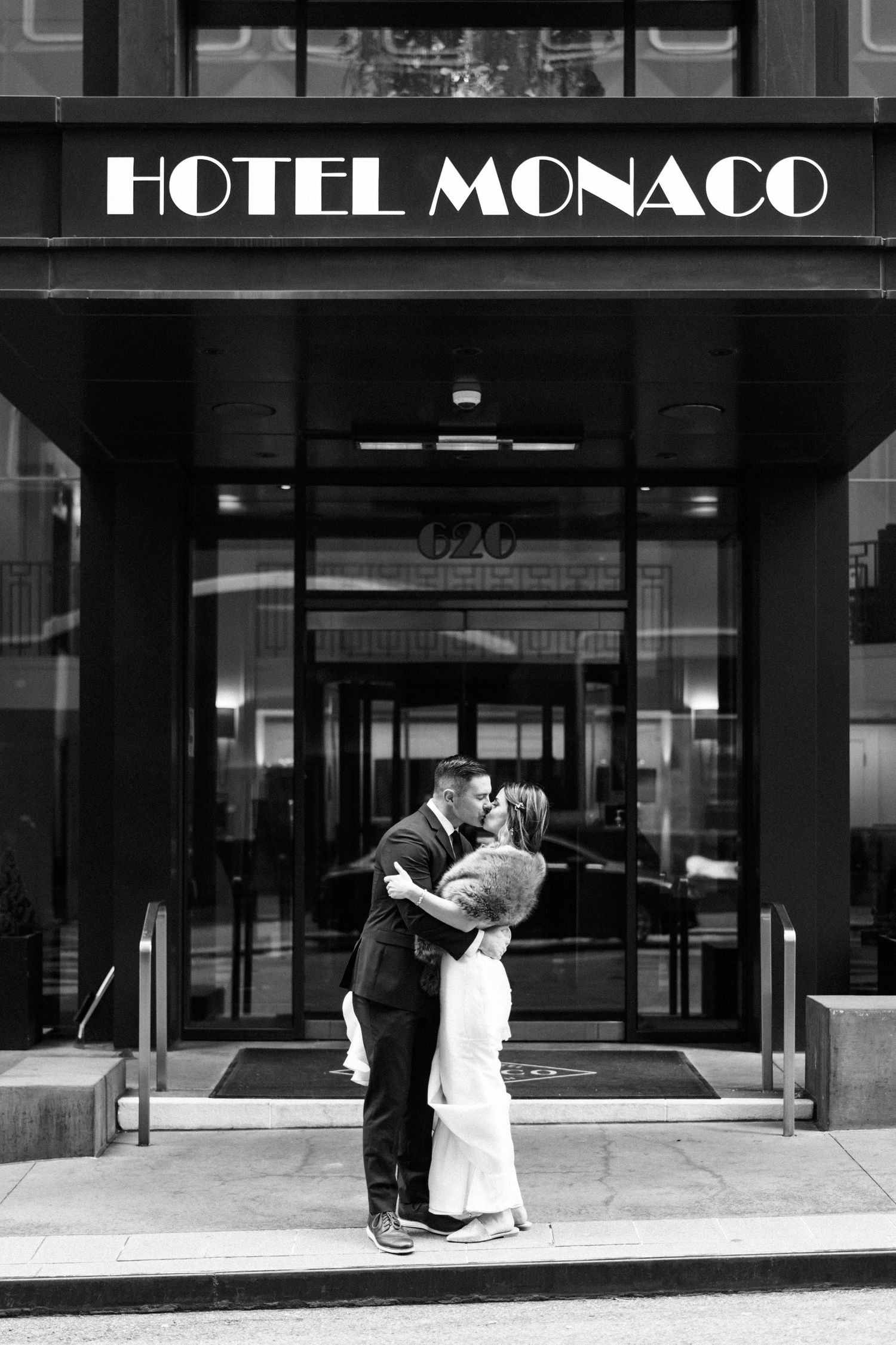 A romantic couple embraces in front of the Hotel Monaco entrance in a classic black and white photograph.
