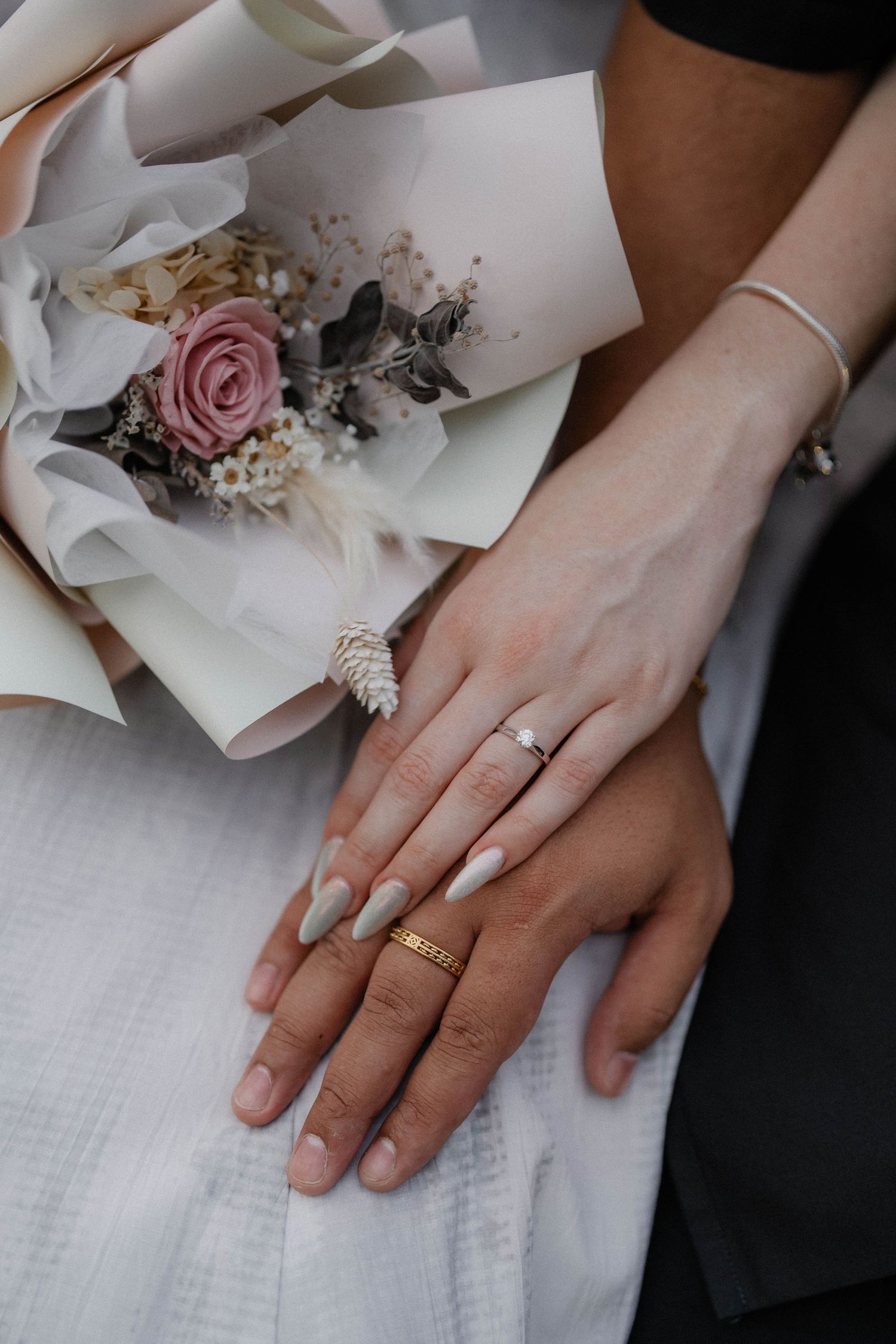 Close-up of hands wearing wedding rings next to a delicate pink and white floral bouquet wrapped in paper.