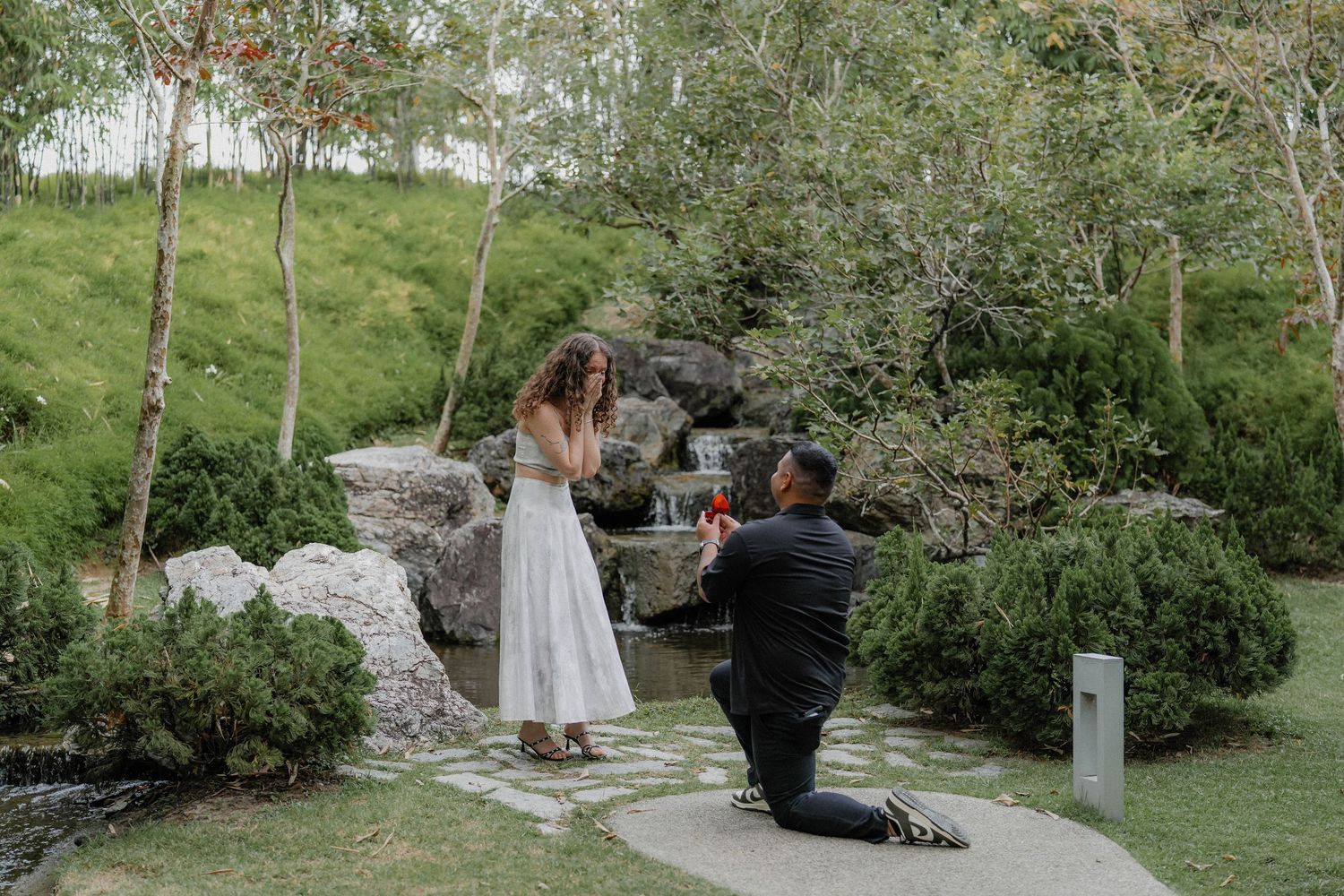A proposal scene by a scenic waterfall in a lush garden setting.