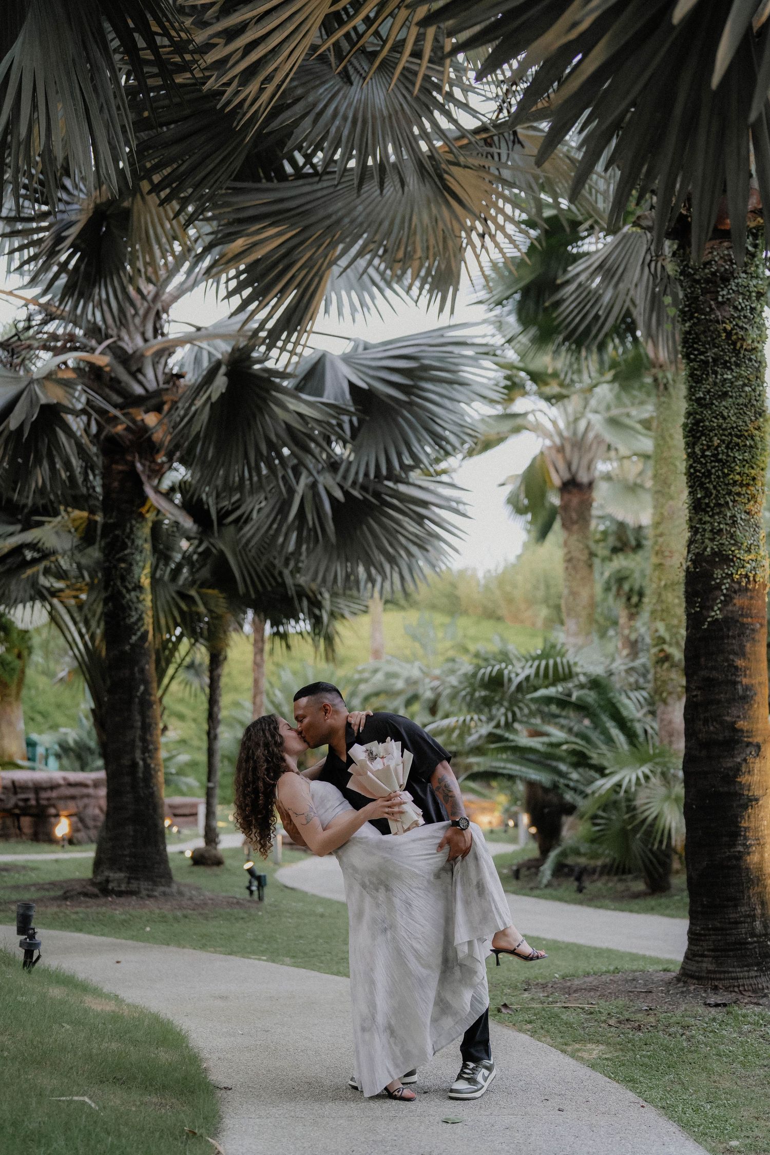 A couple embraces romantically under palm trees in a tropical garden setting.