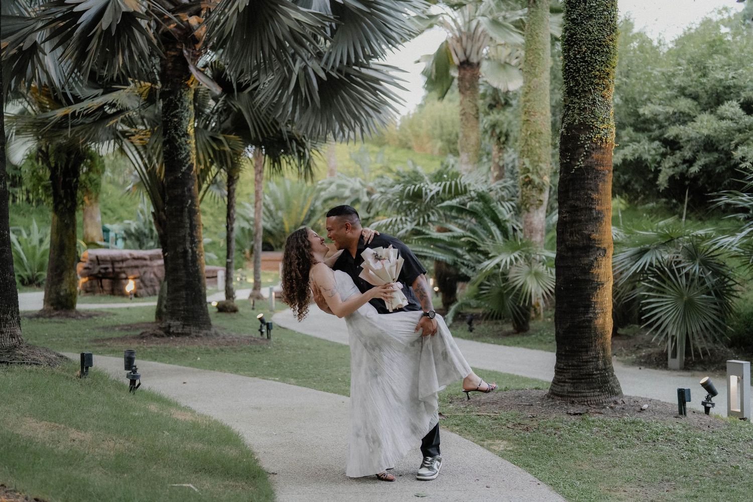 A romantic moment among palm trees in a tropical garden setting with a couple embracing on a pathway.