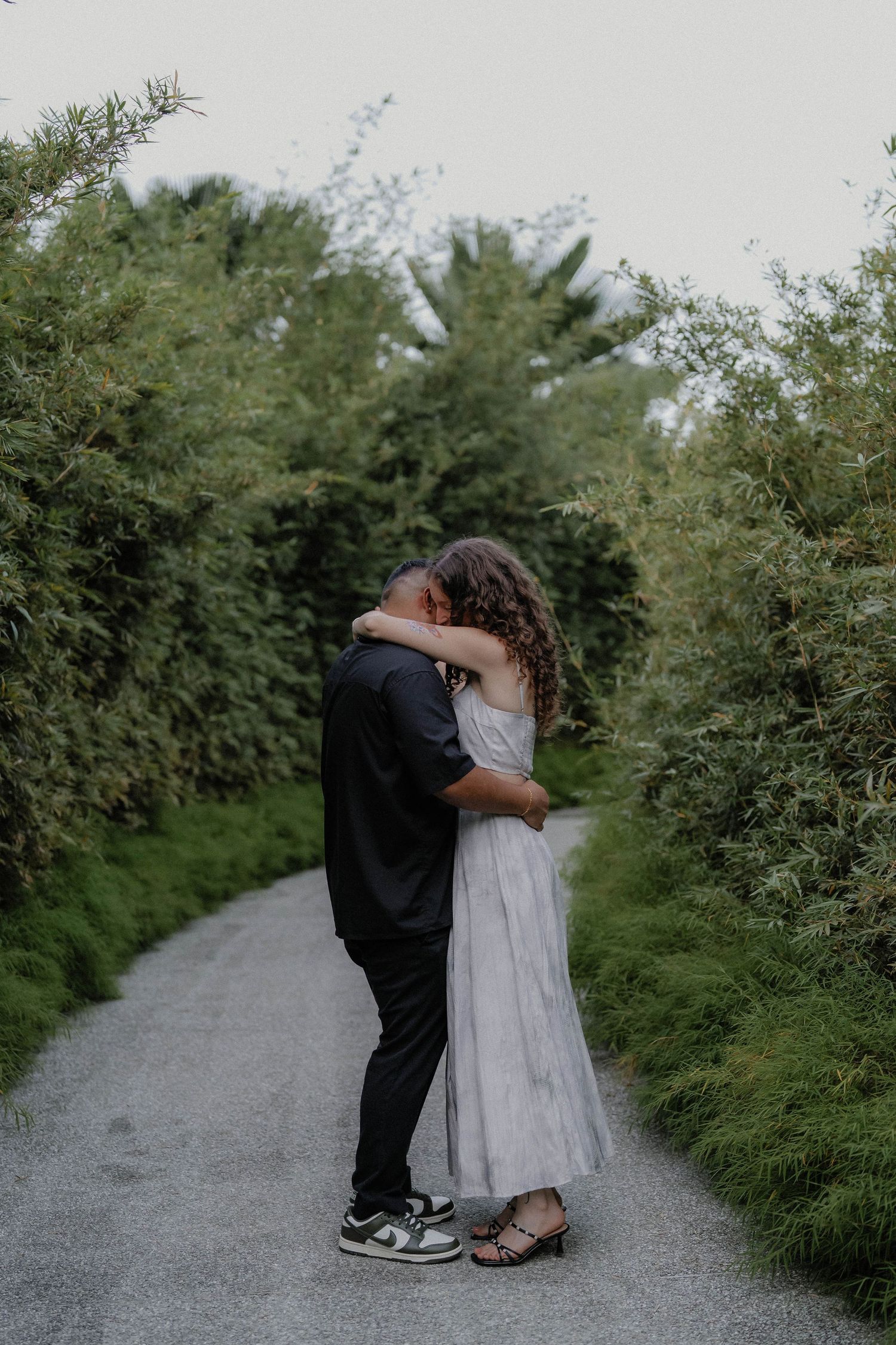 Two people share an intimate moment on a gravel path surrounded by lush greenery.