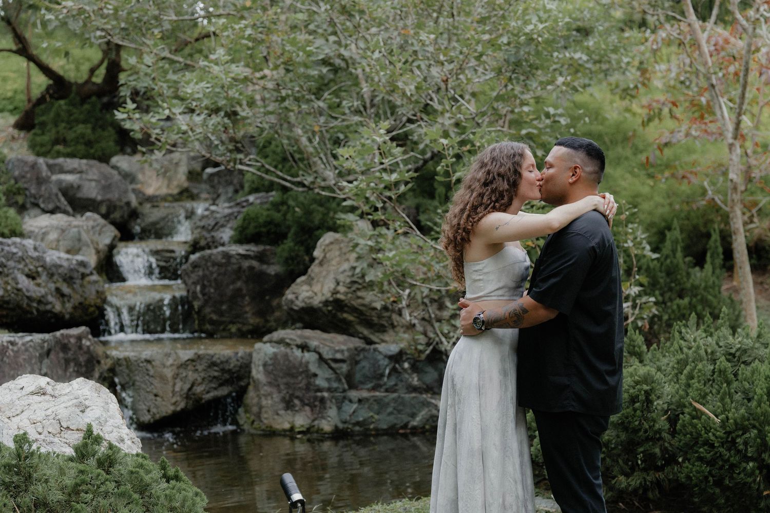 Tender moments shared near a rocky waterfall surrounded by tropical foliage.