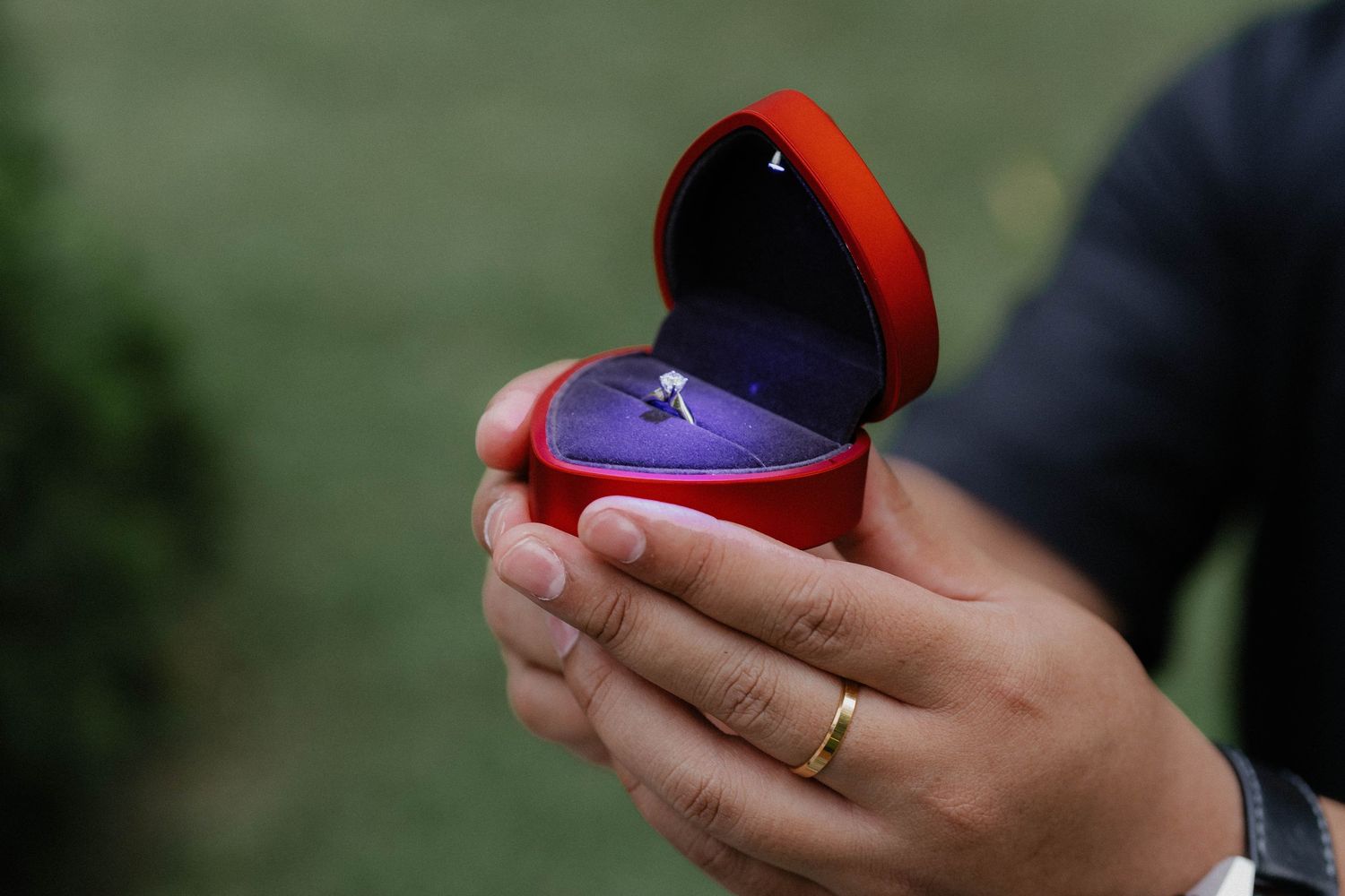 A close-up of hands holding a red heart-shaped ring box with an engagement ring inside.
