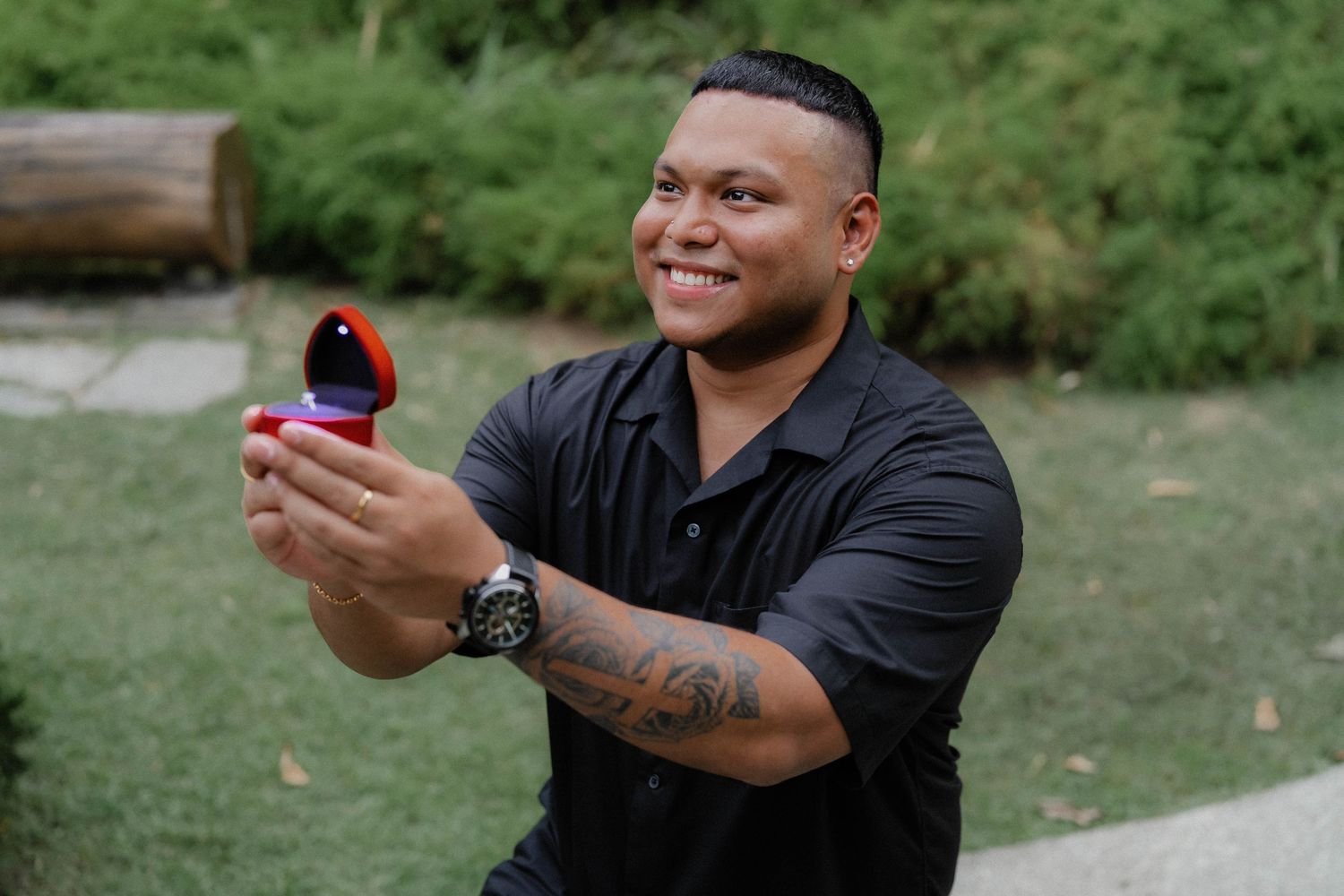 Person in black shirt kneeling while presenting a small box in an outdoor setting.