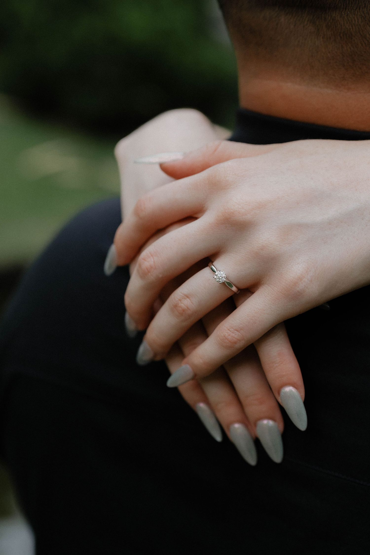 Close-up of elegant hands with a sparkling engagement ring and neutral manicured nails against a dark background.