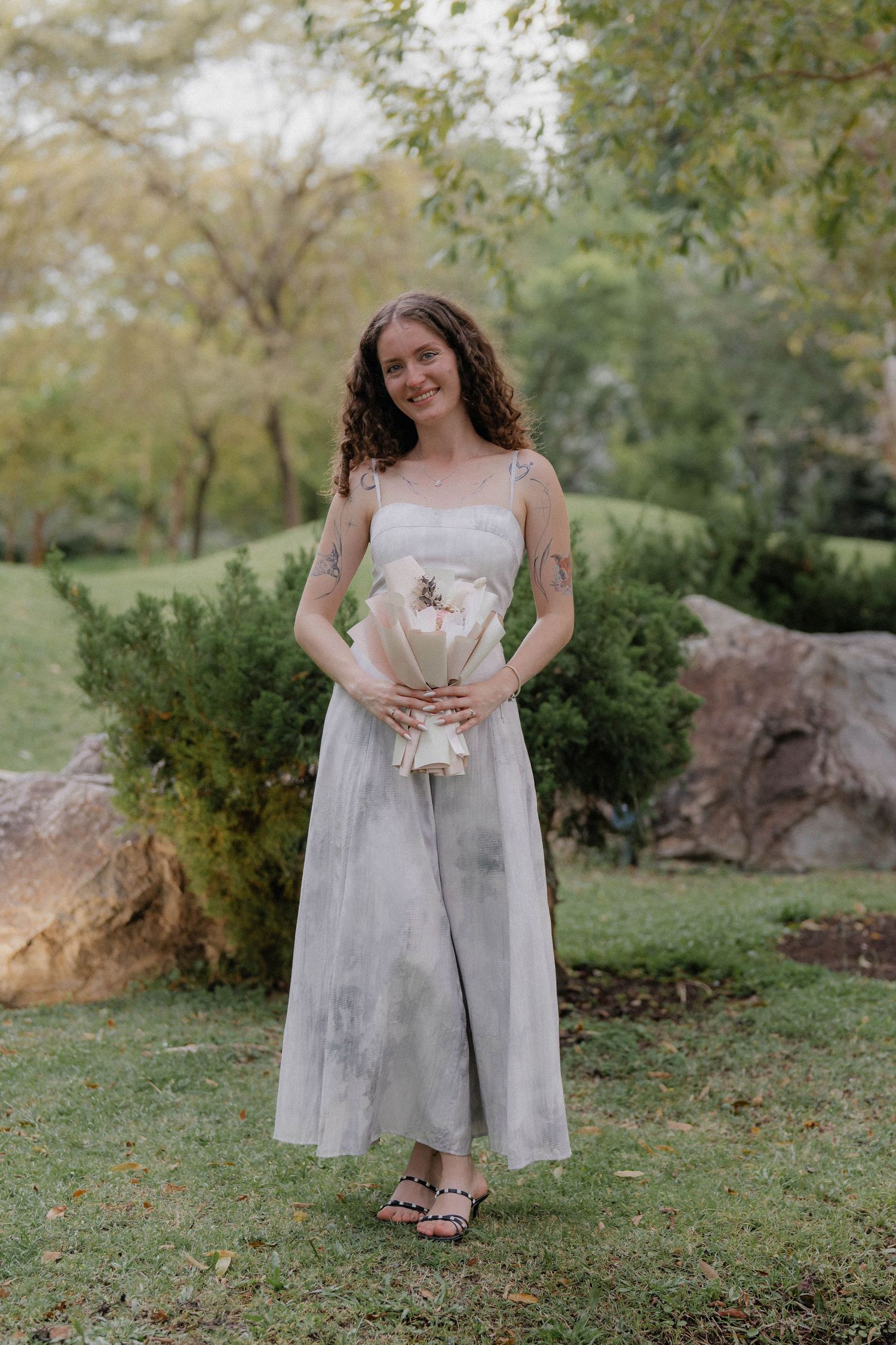 A woman in a light-colored dress holds a bouquet while standing in a garden setting.