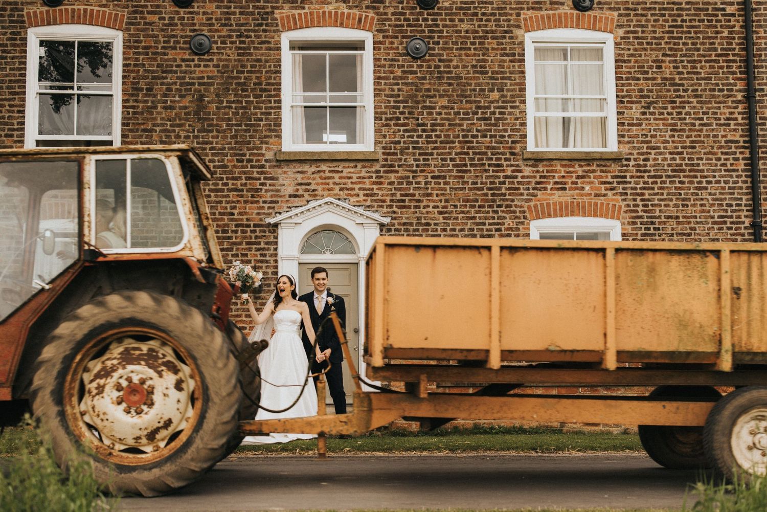 A rustic wedding scene features a couple posing beside a vintage tractor and trailer against a brick farmhouse wall.