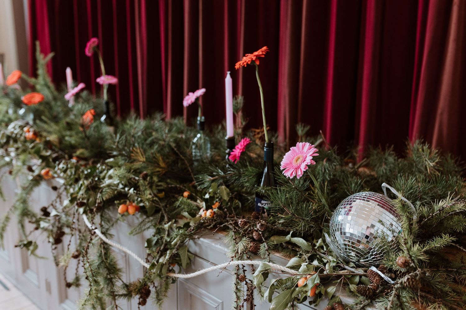 Holiday decorations with pink gerbera daisies, evergreen branches, and a silver disco ball against red velvet drapes.
