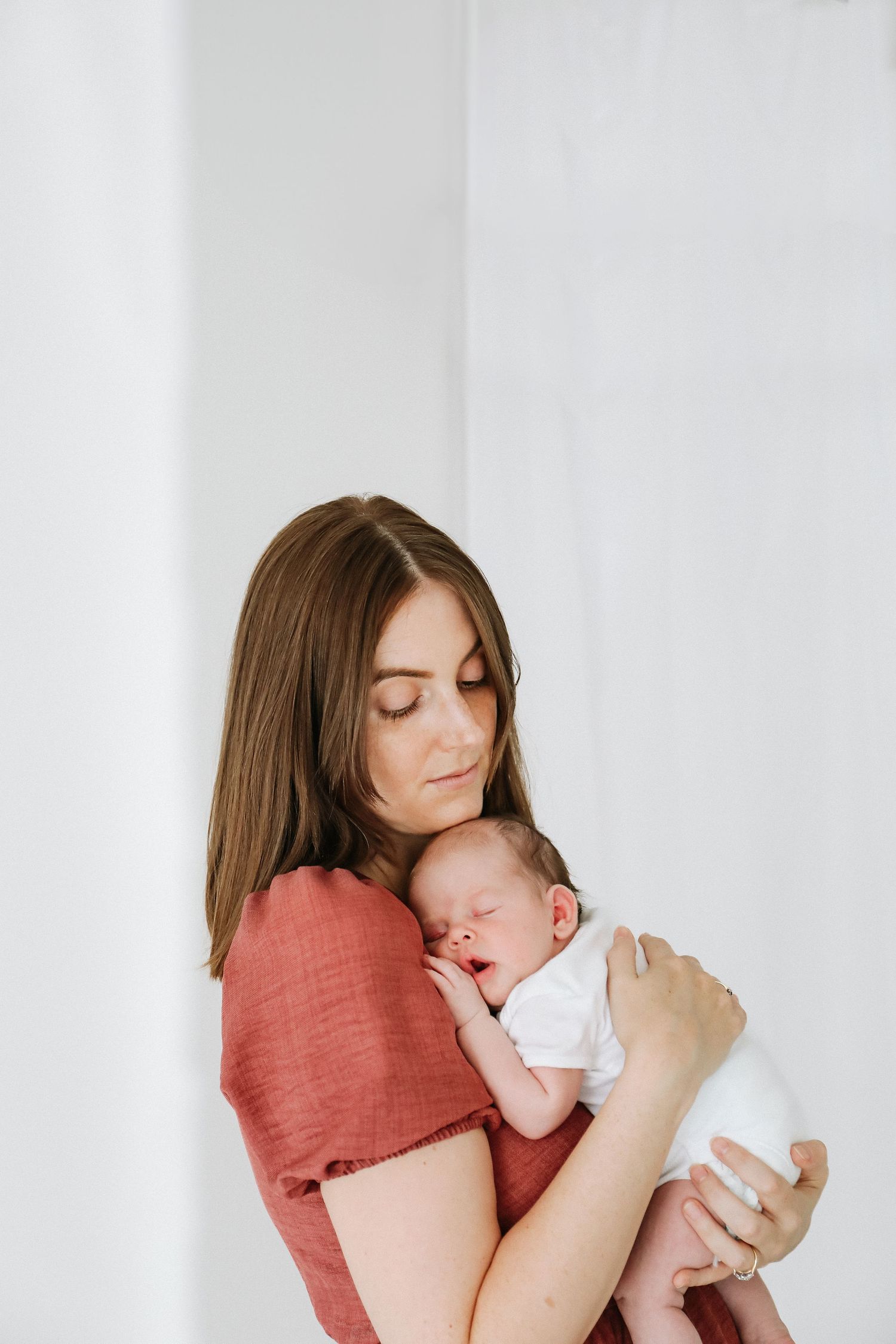Mother in red dress holding tiny sleeping newborn in white romper, natural studio portrait, during newborn photoshoot in Nottingham