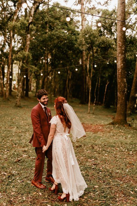 Couple in burgundy suit and lace dress pose in woodland setting with string lights overhead.