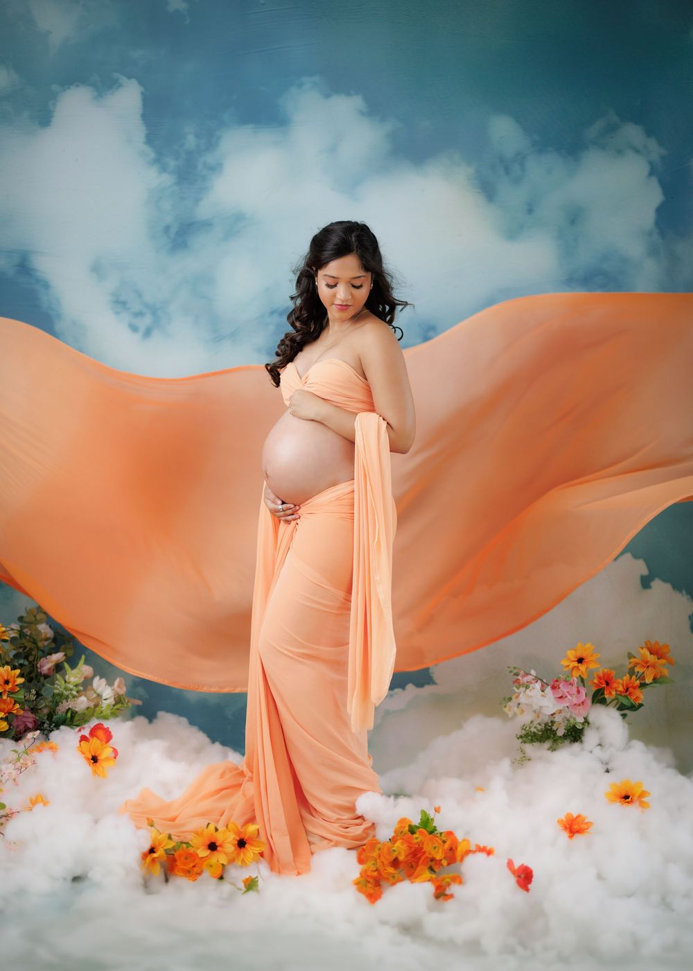 A dreamy maternity photoshoot features a flowing peach gown with orange flowers and wispy clouds in a teal backdrop.
