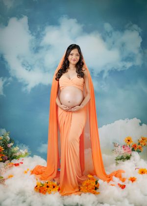 A maternity portrait with someone wearing a flowing peach gown surrounded by orange flowers against a cloudy blue backdrop.