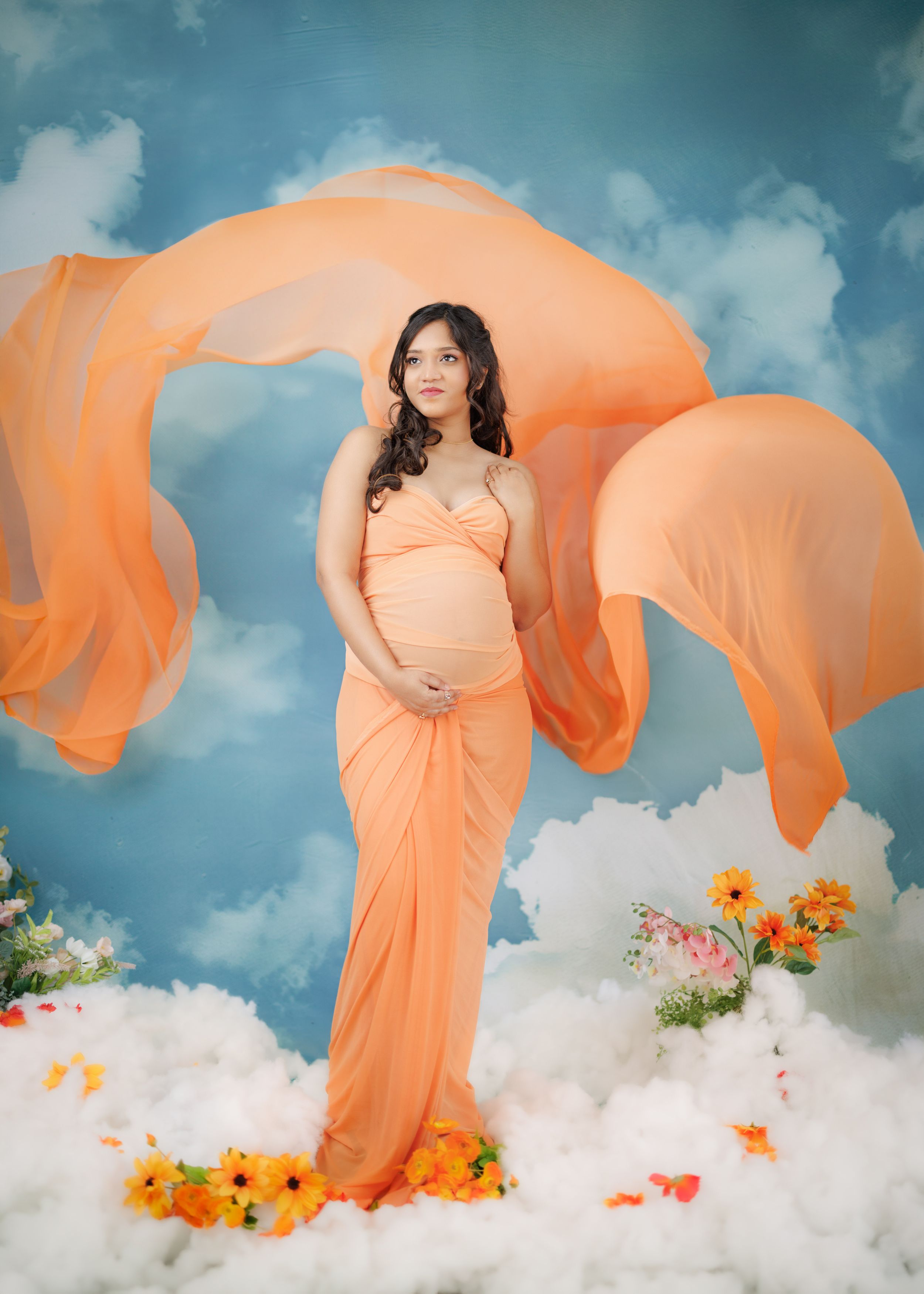 A maternity photo shoot with a flowing peach dress against a dreamy cloud backdrop with orange and yellow flowers.