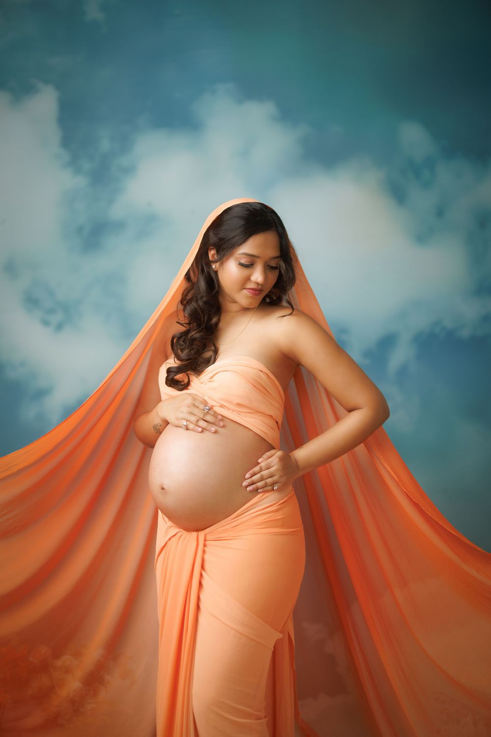 A maternity photoshoot features a flowing orange dress against a moody blue sky background in multiple artistic poses.