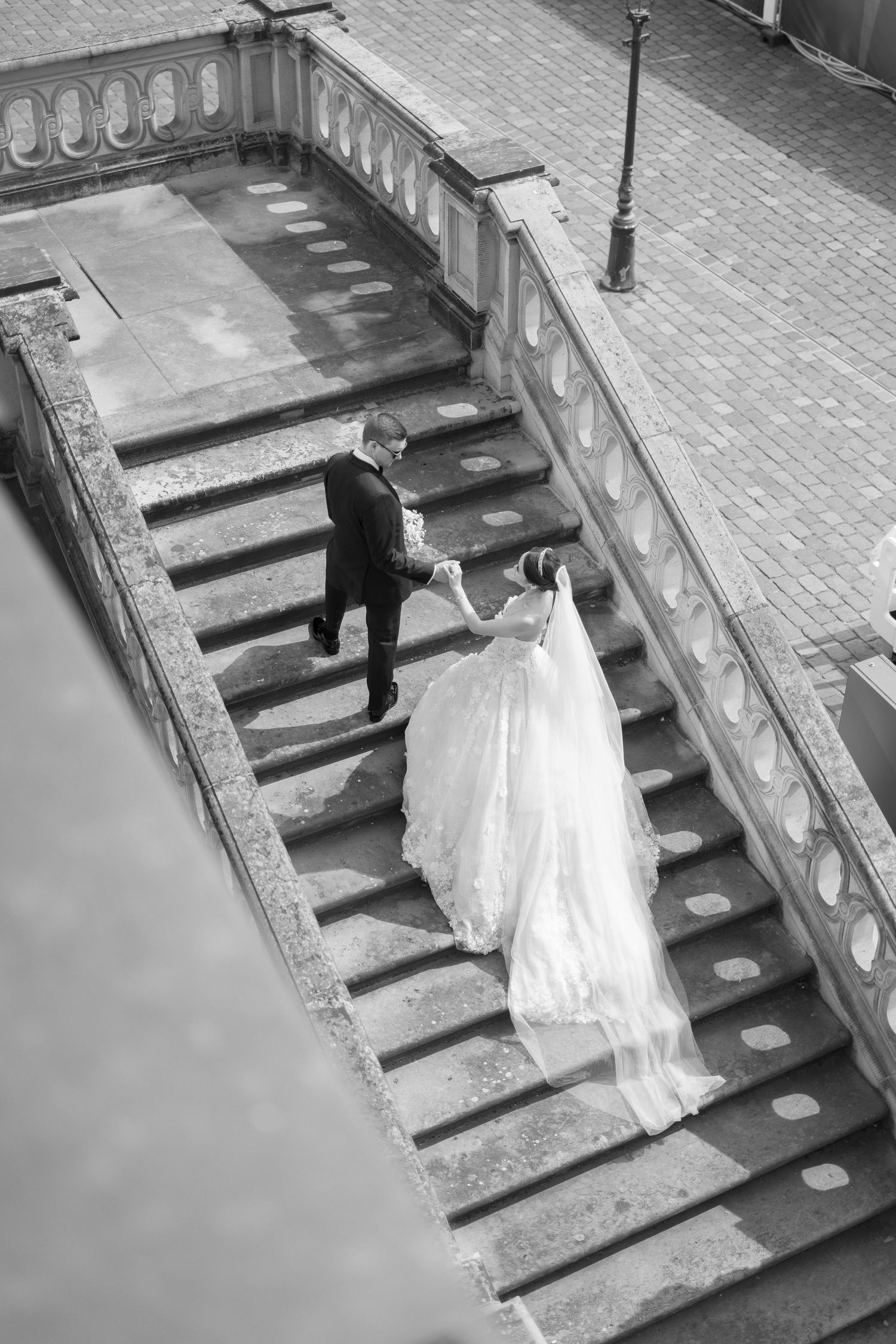 A couple walking down ornate stone steps, with a flowing white wedding dress trailing behind them in black and white.