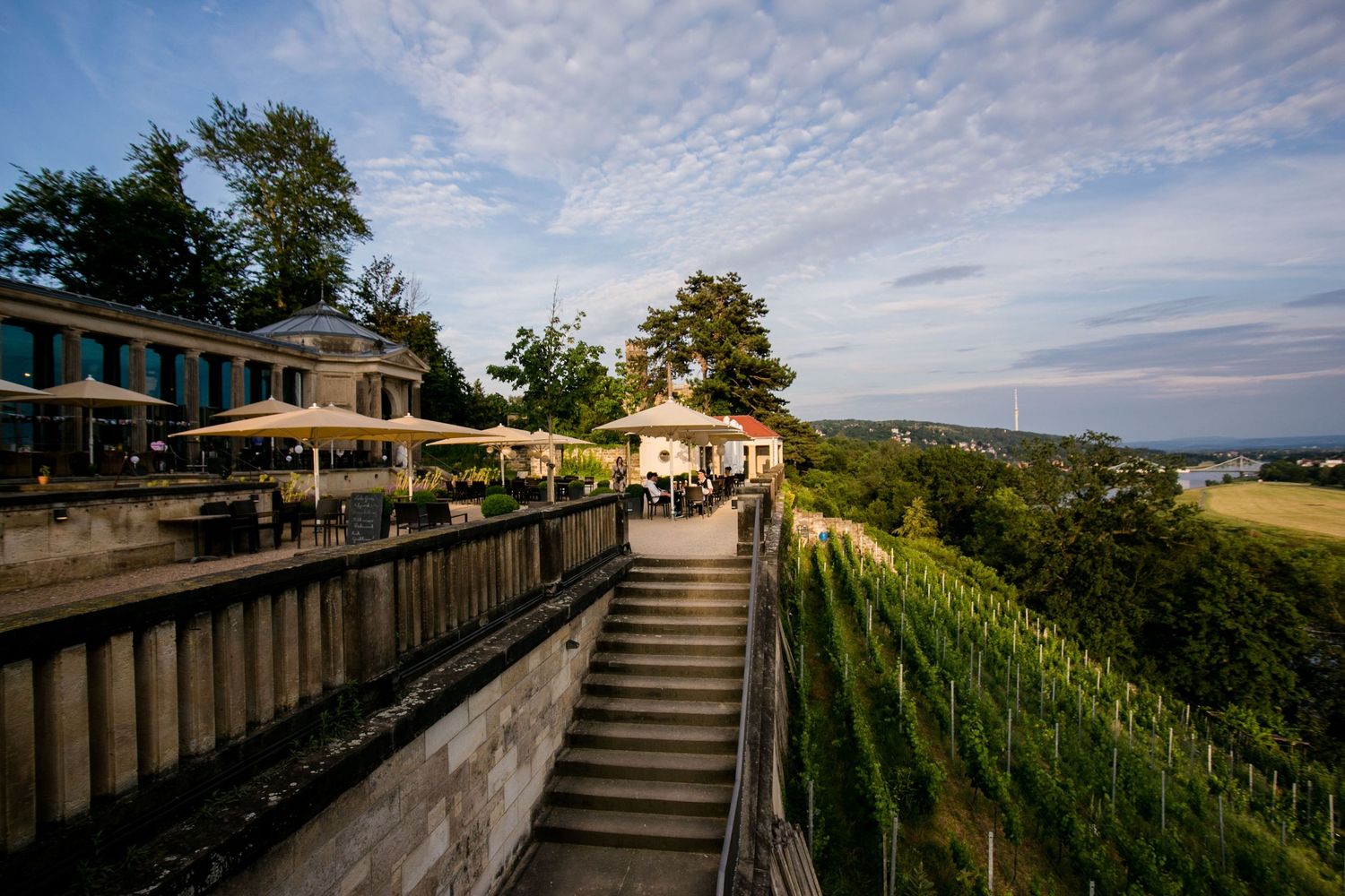 A hillside terrace with stairs and outdoor dining area overlooking vineyards at sunset with scattered clouds above.