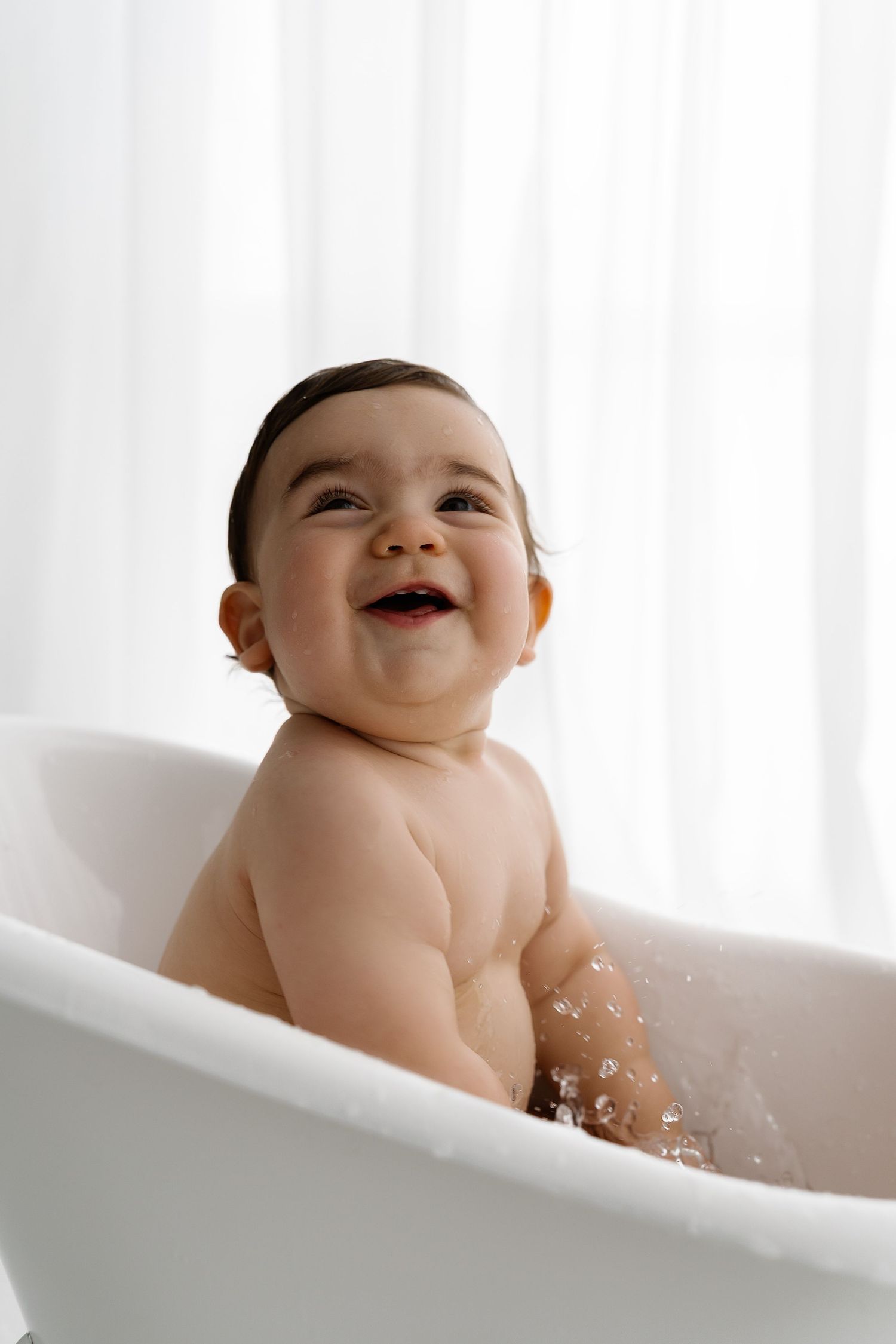 A smiling baby enjoys splashing during bathtime in a white tub.