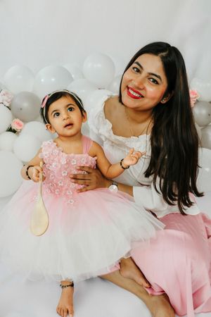Mother and baby daughter in matching pink and white dresses pose against balloon backdrop during celebration photoshoot.