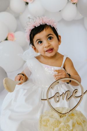 A small child in a white dress with a pink crown holds a wooden 'one' sign against white balloon backdrop.
