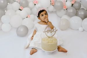 Baby sits in festive party setting with gray and pink balloon arch backdrop and white decorative elements.