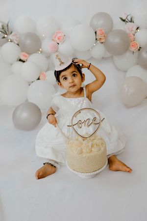 Baby in white lace dress sits by cake with balloons at first birthday celebration with gray and pink decor.