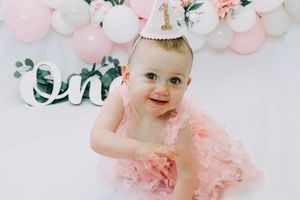 Baby in pink tulle dress sits among soft pink and white balloons and floral decorations for a birthday celebration.