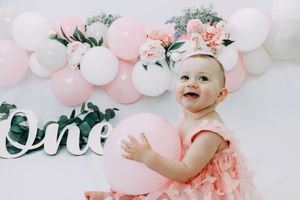 First birthday celebration setup with pink and white balloon arch, floral decorations and silver 'One' text display.