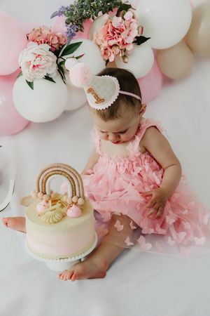 Baby in pink tulle dress celebrates with birthday cake decorated with rainbow arch amid pink and white balloon decor.