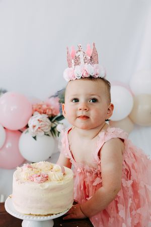 Baby in pink tulle dress and crown sits next to white birthday cake with pink balloons in background.