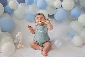 Baby wearing turquoise romper poses under blue and white balloon arch decorations.