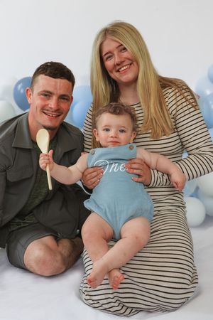 Parents and baby laying together on floor with blue and white balloons for a first birthday photoshoot.