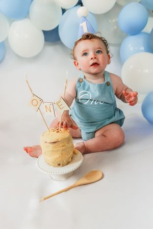 Baby in blue outfit reaching for birthday cake with white and blue balloon backdrop.