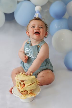 A happy baby in a blue romper sits with a messy cake during a festive first birthday photoshoot with blue balloons.