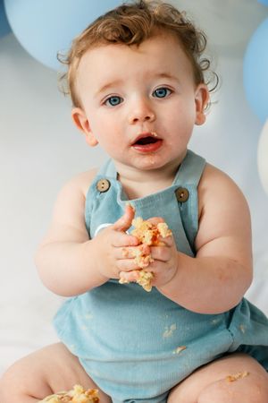 A small child in light blue overalls holds onto messy food with both hands.