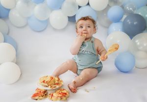 Baby in blue outfit sits among blue and white balloons while messily enjoying smash cake for first birthday celebration.
