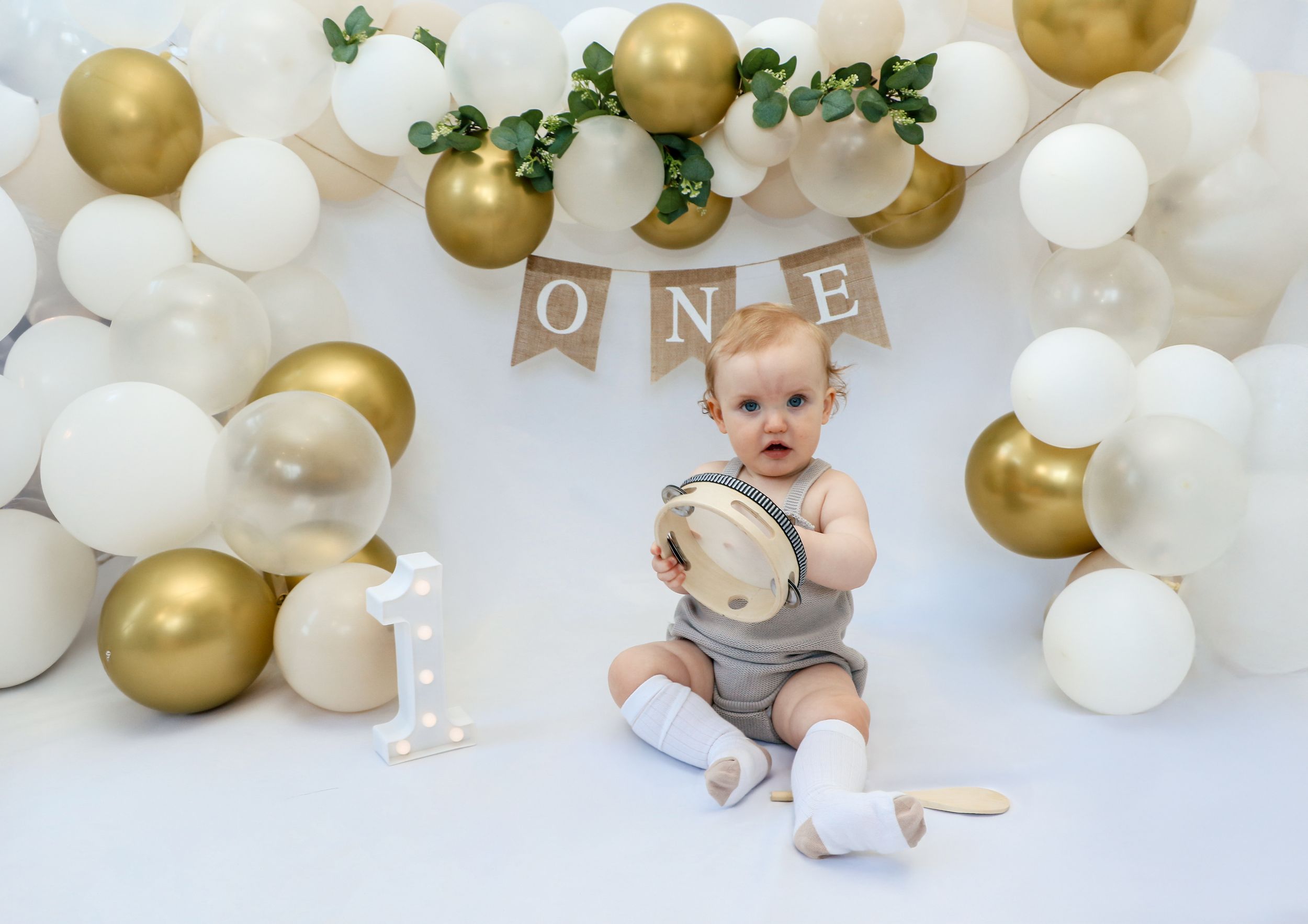 A baby celebrates their first birthday with a backdrop of white and gold balloons and greenery decorations.
