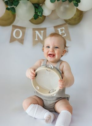 Baby in gray outfit sits under balloon decor holding tambourine during first birthday photoshoot.