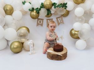 First birthday cake smash photo setup with gold and white balloons and rustic wood slice cake stand against white backdrop.