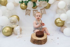 First birthday cake smash photo setup with white and gold balloons, rustic wood slice, and banner decoration.